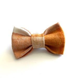 May include: A bow tie featuring a diagonal twill pattern in shades of burnt orange and cream. The fabric has a textured appearance with a central knot that secures the two pleated sides, creating a structured and symmetrical accessory design.