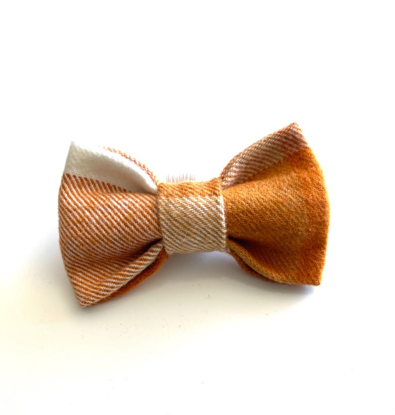 May include: A bow tie featuring a diagonal twill pattern in shades of burnt orange and cream. The fabric has a textured appearance with a central knot that secures the two pleated sides, creating a structured and symmetrical accessory design.