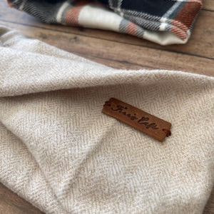 May include: A beige herringbone fabric pouch featuring a brown leather tag embossed with the text "Frari's Cafe". In the background, a second pouch is visible, showcasing a plaid pattern in shades of orange, black, white, and grey.
