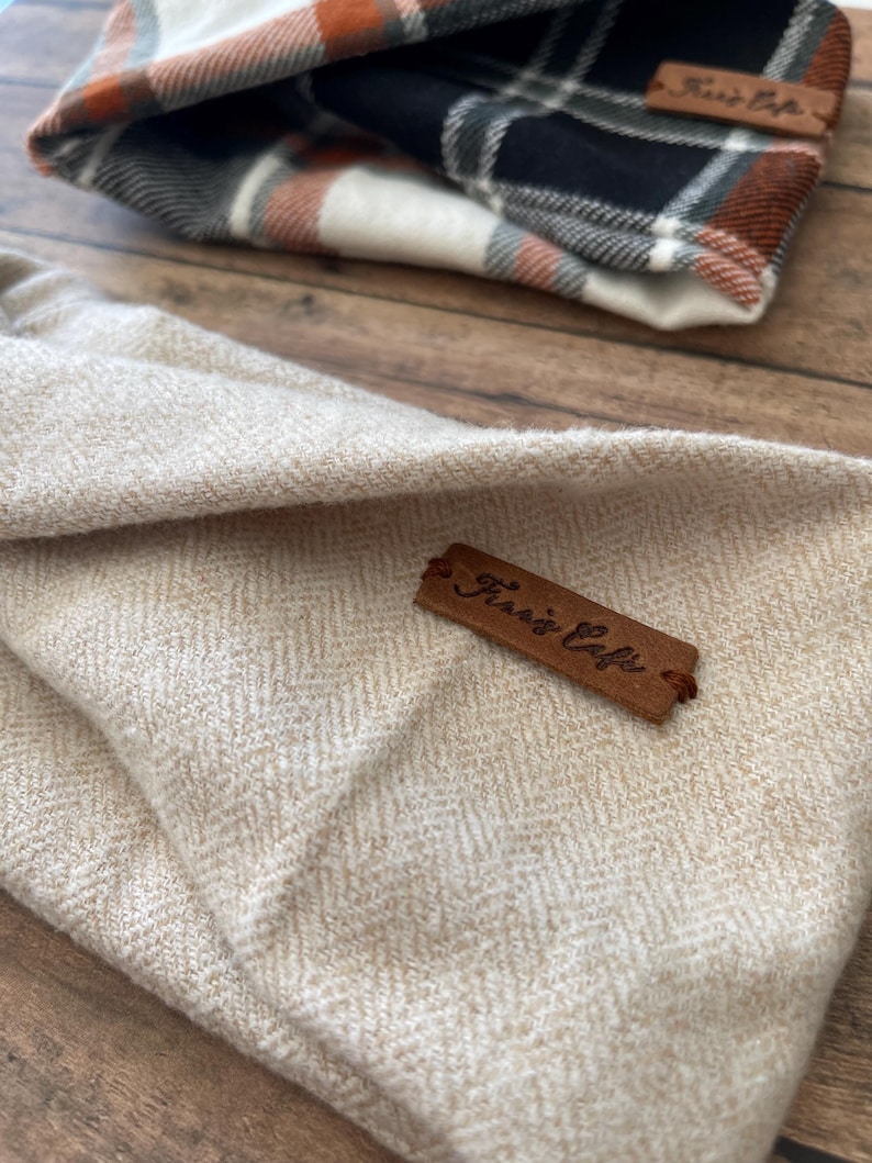 May include: A beige herringbone fabric pouch featuring a brown leather tag embossed with the text "Frari's Cafe". In the background, a second pouch is visible, showcasing a plaid pattern in shades of orange, black, white, and grey.