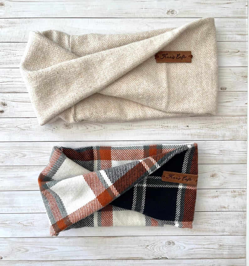 May include: Two fabric headbands with a twisted front design. The top headband is a solid beige with a herringbone texture. The bottom headband features a plaid pattern in orange, white, black, and grey. Both include a small brown leather tag.