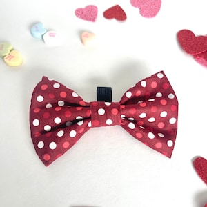 May include: Red and white polka dot bow tie with a black adjustable strap. The bow tie is on a white background with pink and red hearts.