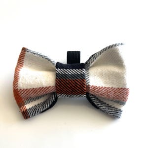 May include: A bow tie featuring a plaid pattern in white, burnt orange, grey, and black. The fabric has a visible woven texture, and the centre knot is wrapped in a matching plaid fabric with a small black elastic loop extending from the top.