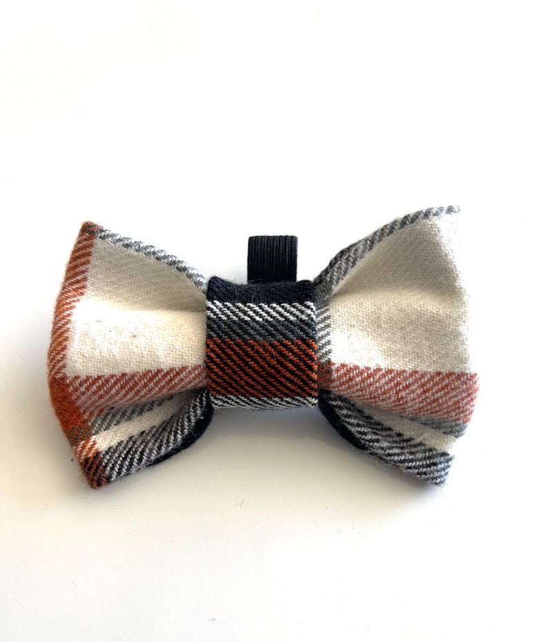 May include: A bow tie featuring a plaid pattern in white, burnt orange, grey, and black. The fabric has a visible woven texture, and the centre knot is wrapped in a matching plaid fabric with a small black elastic loop extending from the top.