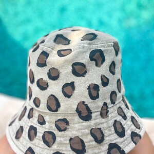 Può includere: Un cappello a secchiello beige con motivo leopardato. Il cappello presenta macchie marroni e nere e un'ampia tesa. Il cappello è realizzato in un tessuto morbido ed è adatto alla protezione solare.