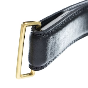 Puede incluir: Un cinturón de cuero negro con una hebilla de metal dorado. La hebilla es de forma rectangular simple con esquinas redondeadas.