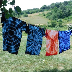 May include: Four tie-dye shirts hanging on a clothesline outdoors. The shirts are blue, blue, orange, and blue. The shirts are hanging in a rural setting with green grass and trees in the background.