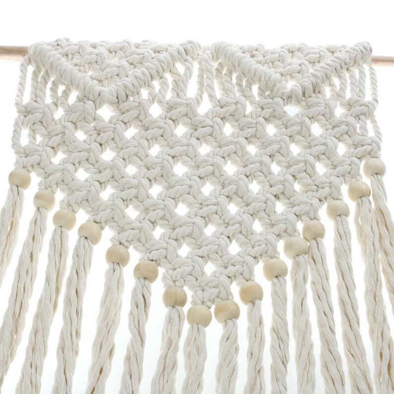 May include: A white macrame wall hanging with wooden beads. The hanging is made of a thick, knotted rope and has a diamond pattern. The beads are evenly spaced along the bottom edge of the hanging.