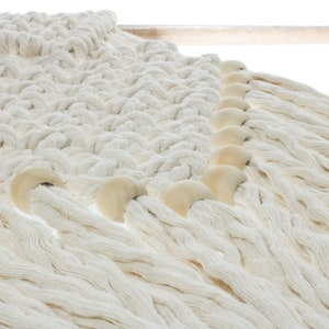 May include: A white macrame wall hanging with wooden beads along the bottom edge. The macrame is made with thick cotton cord and has a fringe along the bottom.