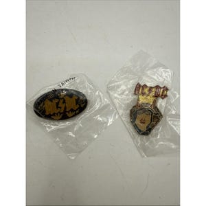 Puede incluir: Dos pines de metal en envases de plástico transparente. Uno es un pin ovalado negro con letras doradas de AC/DC y coronas. El otro es un pin con forma de cara con cuernos, detalles de purpurina dorada y el logo rojo de AC/DC encima.