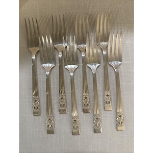 May include: Eight silver forks are displayed. Each fork features a tined head, a decorative handle with an ornate floral design, and a long, slender stem. The forks are arranged on a light-coloured surface.