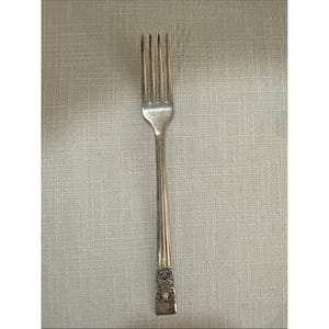 May include: A silver fork with four prongs. The handle features a decorative floral pattern at the base and vertical lines along its length. The fork is made of a shiny, reflective metal.