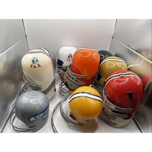 Op de afbeelding: Een verzameling miniatuur American footballhelmen in verschillende kleuren, waaronder rood, oranje, geel en grijs. Elke helm heeft teamlogo's en ontwerpen, met een zilveren gezichtsmasker. De helmen zijn samen gerangschikt en tonen een verscheidenheid aan NFL-teams.