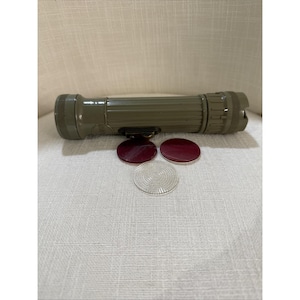 Könnte beinhalten: Eine olivgrüne Militär-Taschenlampe mit geripptem Gehäuse und strukturiertem Ende. Drei runde Linsen, zwei rote und eine klare, sind darunter positioniert. Die Taschenlampe ist für den taktischen Einsatz konzipiert.