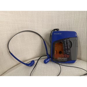 以下が含まれることがあります： 青とグレーのソニーウォークマン スポーツ FM/AMラジオと、それに合う青いヘッドホン。ポータブルカセットプレーヤーには透明な窓とラジオ周波数のダイヤルが付いています。ヘッドホンはグレーのバンドと青いイヤホンが付いています。