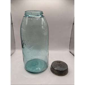 May include: A vintage aqua blue glass Ball Mason jar with a cylindrical body and a threaded neck. The glass features visible air bubbles and embossed lettering on the side. A separate, circular, weathered metal lid sits beside the glass jar.