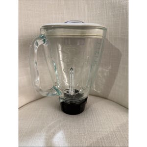 May include: A clear glass blender jar with a white lid and a black base. The jar has a handle and measurement markings. The blender is sitting on a white upholstered chair.