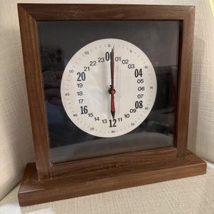 May include: A square wooden clock with a white face and black numbers. The clock has a black background and red hands. The clock is in a dark brown wooden frame and sits on a wooden base.
