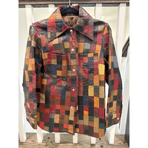 Puede incluir: Una chaqueta de manga larga con botones, confeccionada con un mosaico de cuadrados de cuero multicolor en tonos rojo, marrón, amarillo, azul y negro. La chaqueta presenta un cuello puntiagudo, dos bolsillos en el pecho y botones de presión metálicos.
