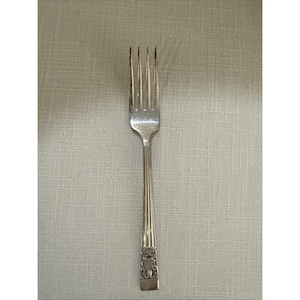 May include: A silver fork with four prongs. The handle features a ribbed design and a decorative floral pattern near the base. The fork is set against a neutral, textured background.