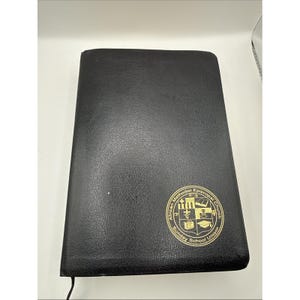 Könnte beinhalten: Schwarzes, ledergebundenes Buch mit einem goldenen Emblem mit den Worten "African Methodist Episcopal Church" und "Sunday School Union". Das Buch ist rechteckig und scheint geschlossen zu sein. Der Einband hat einen leichten Glanz.