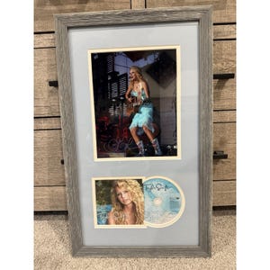 May include: Framed artwork featuring a concert photo and a CD. The top image shows a performer on stage in a blue dress, playing a guitar. The bottom image is a portrait with the artist's name. The CD is also visible.
