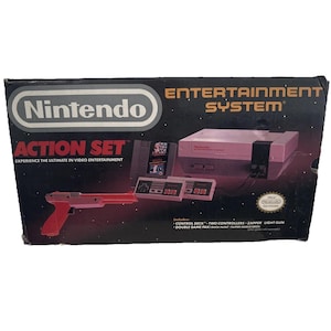 Puede incluir: Caja de un conjunto Nintendo Entertainment System Action Set vintage. La caja presenta una consola, dos mandos, una pistola Zapper y un paquete de dos juegos. La caja es negra con el logotipo de Nintendo y los detalles del producto.
