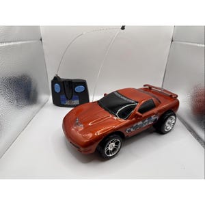 May include: A remote-controlled orange sports car with black windows and silver rims. The car has a spoiler and a logo on the side. A black remote control with blue buttons and an antenna is next to the car.