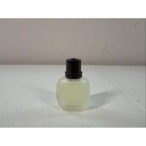 May include: A small, frosted glass bottle with a dark brown cap. The bottle has a rounded shape and appears to contain a light yellow liquid. The bottle has a decorative design on the front.