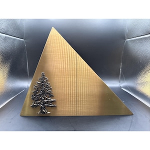 May include: A brass-colored, triangular decorative piece with a textured surface. A detailed, dark-colored tree is affixed to the front. The piece is displayed against a dark background.