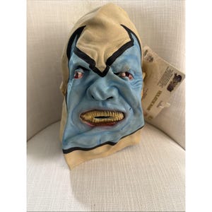 Peut inclure: Un masque d'Halloween en latex avec un visage bleu, une tête jaune et des accents noirs. Le masque a une expression menaçante avec des dents exposées et des yeux rouges. Une étiquette est attachée au masque.