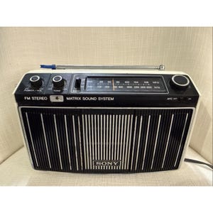 Puede incluir: Radio estéreo FM Sony vintage con un diseño en blanco y negro. La radio tiene un dial de sintonización, control de volumen y el texto "MATRIX SOUND SYSTEM". La parte frontal tiene rayas blancas verticales y el logotipo de Sony.