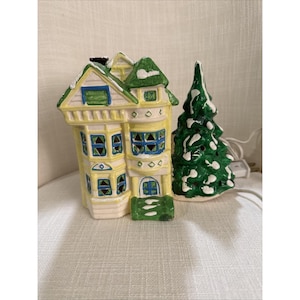 Peut inclure: Une maison et un arbre de village de Noël en céramique. La maison est peinte en jaune avec des accents verts et bleus, et l'arbre est vert avec des détails blancs ressemblant à de la neige. Un cordon blanc s'étend de l'arbre.