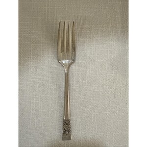 May include: A silver fork with four prongs. The handle features a decorative pattern near the base and a tapered design. The fork is made of a shiny, reflective metal, possibly stainless steel or silver. Suitable for formal dining.