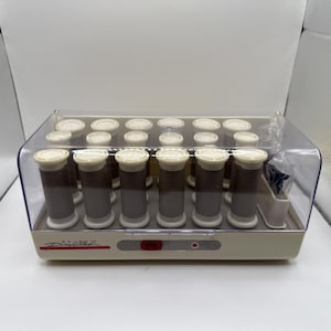 May include: A beige hair roller set with a clear plastic lid. The set includes multiple brown and white rollers of varying sizes. The base has a red power button and a small red light. The brand name is printed on the front.