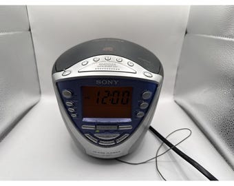 May include: A Sony CD clock radio with a dark gray top, silver accents, and a blue faceplate. The device features a digital display showing 12:00 AM, multiple control buttons, a snooze bar, and a black power cord extending from the right side.