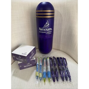 Può includere: Una collezione di articoli di marca viola e bianchi, tra cui un grande contenitore a forma di capsula, blocchi per appunti e penne. Il contenitore ha strisce dorate e il testo "Nexium" con "esomeprazolo magnesio" e "40 mg capsule". Le penne sono in confezioni di plastica.