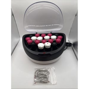 May include: A black and white electric hair setter with a clear hinged lid, containing multiple cylindrical rollers in shades of pink, white, and black. A small clear plastic bag containing several metal hair pins is positioned in front of the unit.