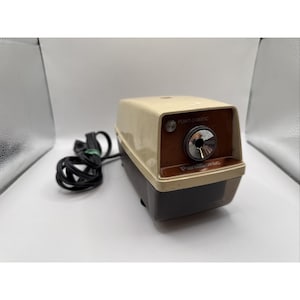 Könnte beinhalten: Ein elektrischer Panasonic Point-O-Matic Bleistiftspitzer mit cremefarbenem Gehäuseoberteil, brauner Frontblende und einem dunkelbraunen, durchscheinenden Spänebehälter. Das Gerät verfügt über ein schwarzes Netzkabel und eine runde Metallöffnung.