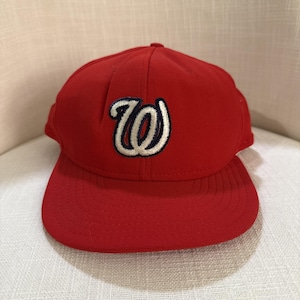 Puede incluir: Gorra de béisbol roja con visera curva. La gorra presenta un logotipo "W" bordado en blanco y azul marino en la parte delantera. El sombrero está hecho de una tela texturizada.