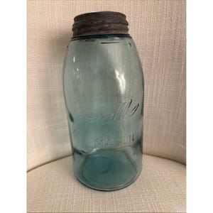 Puede incluir: Un tarro Ball Mason azul vintage con tapa de metal. El tarro tiene letras en relieve que dicen "Ball" y "Mason". El vidrio tiene un ligero tinte aguamarina. El tarro probablemente se utiliza para enlatar o almacenar.