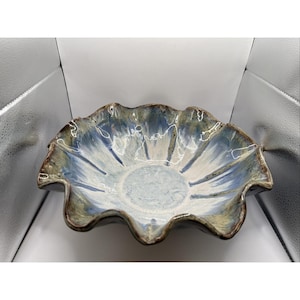 May include: A ceramic bowl with a scalloped, wavy rim featuring a reactive glaze in shades of blue, white, and earthy green. The interior surface displays a radial pattern where the colors blend from a light center toward the dark, textured edges.