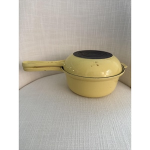 Puede incluir: Una olla amarilla de hierro fundido con tapa negra y asa larga. La olla es redonda con un asa ligeramente curvada. La tapa es plana y tiene un diseño circular. La olla está sobre una silla blanca.