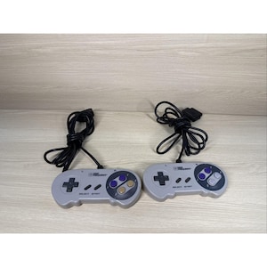 Peut inclure: Deux manettes de Super Nintendo grises avec des câbles noirs. Chaque manette possède un pavé directionnel, quatre boutons d'action et des boutons select/start. Les manettes sont sur une surface en bois clair.
