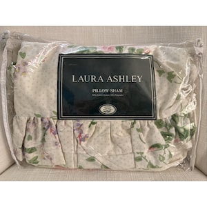 Può includere: Una federa Laura Ashley in una confezione di plastica trasparente. La federa ha uno sfondo bianco con un motivo floreale rosa, viola e verde. La confezione ha un'etichetta nera con le parole "LAURA ASHLEY" e "PILLOW SHAM".