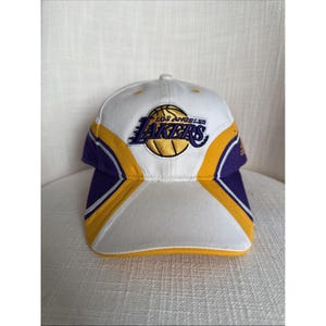 May include: A white, purple, and yellow baseball cap featuring the Los Angeles Lakers logo. The cap has a curved brim and a structured crown. The Lakers logo includes a basketball and the team's name.