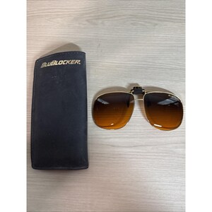 May include: Gold-framed clip-on sunglasses with brown lenses and a black case. The case is rectangular and has the text "BLUBLOCKER" printed on it. The sunglasses are open and ready to be clipped onto a pair of glasses.