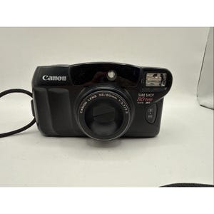 May include: A black Canon Sure Shot 80 Tele Date 35mm film camera featuring a Canon Lens 38/80mm 1:3.7/7.3. The front of the camera displays the brand name, model name, and lens specifications in white and red text, with a built-in flash unit.