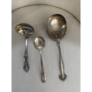Puede incluir: Tres utensilios de servir antiguos de plata: un cucharón, una cuchara y una cuchara de servir. El cucharón y la cuchara de servir tienen mangos ornamentados. La cuchara es más pequeña y tiene un diseño sencillo. Los tres son reflectantes.