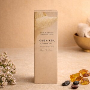 Könnte beinhalten: Eine rechteckige, hellbraune Schachtel für einen Duftdiffusor mit der Aufschrift "God's SPA Harmony". Die Schachtel wird von bernsteinfarbenen Steinen und kleinen, dunkelbraunen Steinen begleitet. Der Text auf der Schachtel lautet "Premium Diffuser Fragrance Sticks" und "100ml / 3.4 fl.oz."